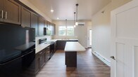 Located at West Division Street in River Falls, Wisconsin, this two-floor residential unit offers a clean, modern design with thoughtfully selected finishes throughout. The open-concept main living area features wood-look luxury vinyl flooring that extends seamlessly from the entry into the kitchen and living spaces, creating a cohesive and easy-to-maintain environment.

The kitchen is a standout feature, equipped with rich dark espresso cabinetry, white countertops, black appliances including a range and microwave, a double-basin stainless steel sink, and a central island with pendant lighting overhead. Recessed lighting and a window allow natural light to fill the space, while a wall-mounted mini-split unit provides efficient climate control.

The bedrooms are carpeted for comfort and include ceiling fans, baseboard heating, and ample natural light from double windows. Closet space is provided within each bedroom. The hallways and connecting areas are also carpeted, contributing to a quiet, residential feel.

The bathroom features dark cabinetry with a white countertop vanity, a large mirror, wood-look flooring, and grab bars, suggesting accessibility-conscious design. Throughout the unit, neutral warm beige tones on the walls, white trim, and panel-style doors maintain a consistent, contemporary aesthetic suited for everyday living.