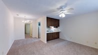 This single-floor space at Summit offers a practical and open layout that connects the kitchen and living areas seamlessly. The kitchen is equipped with dark wood cabinetry providing ample storage, a built-in sink with a countertop that extends into a breakfast bar-style overhang, and a white refrigerator. The cabinetry's rich, dark tones contrast cleanly against the light-colored walls, giving the kitchen a grounded, functional aesthetic.

The living area is generously proportioned, featuring wall-to-wall carpet in a neutral beige tone that carries throughout the space, lending warmth and continuity. A ceiling fan with an integrated light fixture anchors the living area, offering both comfort and illumination. A chandelier is visible further down the hallway, adding a decorative touch to the interior.

The hallway connects the main living space to additional rooms at the rear of the unit, maintaining an easy flow throughout. White textured ceilings and neutral wall tones keep the interior feeling bright and cohesive. Overall, the space presents a clean, no-frills layout well-suited for comfortable everyday living.