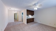 This single-floor space at Summit offers a practical and open layout that connects the kitchen and living areas seamlessly. The kitchen is equipped with dark wood cabinetry providing ample storage, a built-in sink with a countertop that extends into a breakfast bar-style overhang, and a white refrigerator. The cabinetry's rich, dark tones contrast cleanly against the light-colored walls, giving the kitchen a grounded, functional aesthetic.

The living area is generously proportioned, featuring wall-to-wall carpet in a neutral beige tone that carries throughout the space, lending warmth and continuity. A ceiling fan with an integrated light fixture anchors the living area, offering both comfort and illumination. A chandelier is visible further down the hallway, adding a decorative touch to the interior.

The hallway connects the main living space to additional rooms at the rear of the unit, maintaining an easy flow throughout. White textured ceilings and neutral wall tones keep the interior feeling bright and cohesive. Overall, the space presents a clean, no-frills layout well-suited for comfortable everyday living.