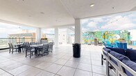 Located atop 920 Manatee Avenue West in Bradenton, Florida, the Nine20 rooftop amenity space is a thoughtfully designed, open-concept retreat that blends covered and open-air environments across a single generously sized floor. The space is defined by clean white architecture, large structural columns, and expansive openings that invite natural light and panoramic views of the Bradenton skyline and surrounding waterways.

The covered interior section features large-format tile flooring, recessed lighting, and wall-mounted flat-screen televisions flanked by speakers, creating a comfortable lounge atmosphere. Plush navy blue sectional seating and sleek low-profile coffee tables are arranged to encourage gathering and relaxation. An indoor dining area provides multiple tables and cross-back chairs for casual meals and social events.

An outdoor kitchen and bar area with professional-grade ventilation hoods and barstool seating serves as a functional entertaining hub, complete with its own mounted television. Adjacent long communal dining tables offer ample seating for group gatherings.

The open-air terrace extends the space further, featuring a fire pit surrounded by vibrant turquoise Adirondack chairs, additional sectional seating, and dining sets with sweeping views protected by glass railings. String lights add a warm ambiance at dusk. A bold, tropical "Nine20" mural anchors the outdoor area, reinforcing the space's vibrant Florida character.