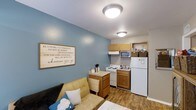 This classic two-bedroom, one-bathroom unit is thoughtfully arranged on a single floor, offering an efficient and comfortable layout. The living area and kitchen share an open-concept space, creating a connected and practical environment ideal for everyday living. The living area features wood-style flooring that adds warmth throughout, complemented by a bold blue accent wall that serves as a focal point of the room. A decorative wall sign adds a personalized, welcoming touch to the space.

The kitchen is compact yet well-equipped, featuring wooden cabinetry, a white gas range, a full-size refrigerator, and a microwave, providing all essential appliances for meal preparation. The kitchen layout is designed to maximize functionality within a modest footprint, with cabinet storage above and below the counter. Flush-mount ceiling light fixtures illuminate both the living and kitchen areas, ensuring consistent, even lighting throughout.

The unit's straightforward single-floor design ensures easy accessibility, while the cohesive color palette and warm flooring create an inviting ambiance suited to comfortable, practical living.