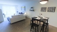 This single-floor residence features an open-concept layout that seamlessly integrates the living and dining areas into one cohesive space. The living area is anchored by a large white sofa accented with blue and purple decorative pillows, complemented by a tall floor lamp and a framed artwork on the wall. Floating shelves above display small decorative items, adding a personalized touch to the clean, neutral-toned walls. Natural light filters in through a window dressed with blinds, brightening the space alongside warm overhead and accent lighting. The dining area is furnished with a dark, high-top table paired with matching bar-height stools, set with glassware and linens, creating an inviting atmosphere for shared meals. A statement pendant light hangs above, adding a stylish focal point. Wall décor, including a bold typographic sign, contributes to the warm and communal ambiance. Neutral-toned carpeting runs throughout, tying both areas together cohesively. The overall design balances comfort and functionality, making it a practical yet welcoming living environment.