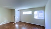 This single-floor space presents a clean, functional layout with a welcoming, understated aesthetic. The main living area is finished with rich hardwood flooring that runs throughout, adding warmth and visual continuity to the space. Neutral beige walls provide a versatile backdrop suitable for a variety of furnishing styles.

Natural light is a notable feature of the room, with two horizontal windows fitted with white blinds positioned along the main wall. These windows allow daylight to filter in, casting bright patches across the hardwood floor and contributing to an open, airy feel despite the modest scale of the space.

A white entry door is positioned to one side of the room, accompanied by what appears to be a utility panel mounted on the adjacent wall. An HVAC vent is visible near the ceiling, indicating a central climate control system. White baseboards frame the perimeter of the room, providing a crisp contrast to the darker flooring.

Overall, the space maintains a straightforward, practical character — well-lit, easy to maintain, and adaptable for a range of uses. The combination of hardwood floors, neutral tones, and natural light creates a bright and functional environment.