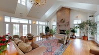 This spacious common lounge is defined by its dramatic vaulted ceilings that rise to an impressive height, creating an open and airy atmosphere throughout. A striking floor-to-ceiling brick fireplace serves as the room's centerpiece, topped with a decorative wreath and flanked by candle sconces, lending a warm, traditional character to the space. An elegant gold chandelier hangs prominently, complementing the room's classic aesthetic.

Natural light floods the interior through multiple sets of white-framed French doors and large divided-light windows positioned along the perimeter, offering views of the surrounding landscape and enhancing the bright, welcoming ambiance. The warm hardwood flooring anchors the space and is partially covered by a large, richly patterned area rug in traditional motifs of red, blue, and ivory.

The lounge is furnished with comfortable upholstered seating arrangements, including sofas, armchairs, and dining-height chairs grouped to encourage both relaxed conversation and small gatherings. Lush potted greenery is thoughtfully placed throughout, adding a natural, refreshing element. Wall sconces provide supplemental lighting, contributing to the overall inviting and refined character of this communal gathering space.