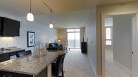 This single-floor residence showcases an open-concept design that seamlessly connects the kitchen and living areas. The kitchen features a granite-topped breakfast bar with seating for two black cushioned bar stools, complemented by pendant lighting and a track lighting system overhead that illuminates the space warmly.

The living area is finished with neutral wall-to-wall carpeting and light gray walls, creating a clean and cohesive aesthetic. A dark sofa adorned with blue accent pillows anchors the seating area, which is centered around a coffee table and paired with a media console. A wall-mounted television is positioned nearby for comfortable viewing.

The standout feature of the living space is a large floor-to-ceiling window that floods the room with natural light and provides direct access to a private balcony, where outdoor seating is visible. The expansive window frames an open exterior view, adding a sense of airiness to the interior.

Decorative elements, including candle holders and framed wall art, contribute to the overall welcoming ambiance of this well-appointed space.