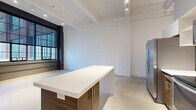 This industrial-style loft showcases a striking blend of raw architectural character and modern finishes across a single floor. The open living and kitchen area anchors the space, featuring polished concrete floors, exposed structural beams, and a sleek kitchen island with wood-toned cabinetry and a stainless steel refrigerator. A row of large, dark-framed factory windows floods the room with natural light while offering expansive views, and a track lighting system runs along the ceiling to complement the industrial aesthetic. An exposed HVAC duct adds to the loft's urban character.

A long hallway connects the main living area to the private rooms, flanked by a painted brick wall that reinforces the building's industrial origins. Both bedrooms are finished with dark textured carpet and crisp white walls, providing a calm contrast to the open living space. The primary bedroom includes a sliding door closet with built-in shelving for organized storage. The laundry room is equipped with a full-size washer and dryer, adding everyday convenience. Polished concrete continues through the hallway and laundry area, maintaining a cohesive aesthetic throughout. The space offers a functional, well-considered layout with a distinctly urban loft ambiance.