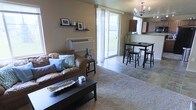This two-floor space offers a thoughtfully arranged open-concept layout on the main level, where the living area flows seamlessly into the dining and kitchen spaces. The living area is anchored by a warm brown leather sofa accented with teal and patterned throw pillows, set atop a plush area rug. A wall-mounted shelf displays framed artwork and decorative accents, adding a personal touch to the neutral-toned walls. A sliding glass door floods the space with natural light and provides direct views of the surrounding greenery, creating a bright and welcoming atmosphere. A wall-mounted HVAC unit is conveniently positioned nearby.

The dining area features a dark wood high-top table surrounded by matching bar-height stools, illuminated overhead by an elegant chandelier that adds a refined focal point to the space. The tiled flooring in the dining and kitchen zones contrasts tastefully with the carpeted living area. The kitchen is equipped with dark upper cabinetry and appears to open toward the dining space, supporting a cohesive and functional layout ideal for everyday living and entertaining.