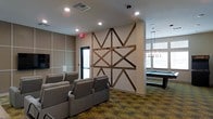 This single-floor amenity lounge is thoughtfully designed to offer residents a versatile space for leisure and socializing. The room is immediately distinguished by a large, geometric wood accent wall crafted from rustic barn-style timber, forming an eye-catching star or compass pattern against a neutral beige backdrop. This architectural focal point bridges contemporary and rustic design sensibilities, lending warmth and character to the space.

The lounge is divided into distinct activity zones. A media area features rows of plush, gray upholstered recliner-style seating arranged to face a wall-mounted flat-screen television, creating a comfortable theater-like atmosphere. Adjacent to this seating area, a billiards table sits beneath a sleek, linear multi-bulb pendant light fixture, providing dedicated task lighting and defining that zone of the room.

Recessed ceiling lighting illuminates the space throughout, complementing the natural light that filters in through windows along the perimeter. The patterned carpet in earthy tones of green and gold adds visual texture and cohesion to the overall design. The combination of entertainment features and thoughtful décor makes this lounge an inviting communal destination.