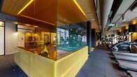 This contemporary fitness center delivers a well-appointed, multi-zone workout environment defined by striking design details and high-quality equipment throughout. The space is organized into distinct functional areas, each thoughtfully separated yet visually connected. A glass-enclosed cycling studio with a perforated yellow metal base serves as a bold architectural centerpiece, bathed in warm amber accent lighting along its ceiling edge and visible from multiple vantage points across the floor.

The cardio zone features a row of treadmills and elliptical machines positioned directly in front of expansive floor-to-ceiling windows, providing unobstructed outdoor views and generous natural light during workouts. Adjacent to this, a dedicated strength area offers a full rack of dumbbells, adjustable benches, barbells, and medicine balls set against textured walls with large mirrors that visually expand the space.

A welcoming entry lounge area features warm wood flooring, a built-in padded bench, and vibrant yellow accent stools beneath a vaulted wood-panel ceiling with recessed lighting. Bold monochromatic dot-patterned wallcoverings add a playful, energetic aesthetic to the darker-toned walls. Rubber flooring throughout the workout zones ensures safety and functionality. The overall ambiance is dynamic and motivating, blending industrial-modern design elements with warm wood tones and bold yellow accents.
