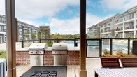 The Pool Grill Area is a well-appointed outdoor amenity space designed for relaxation and entertaining. Sheltered beneath a covered pavilion with sturdy structural columns and an exposed beam ceiling, the area offers an inviting retreat with open views of the surrounding community and pool.

At the heart of the grilling station, two stainless steel gas grills are mounted on handsome brick bases, providing ample cooking capacity for gatherings. The stone tile flooring extends throughout, lending a clean and polished aesthetic to the outdoor setting. Adjacent to the grill stations, a striking metallic-paneled outdoor fireplace serves as a bold focal point, framed by vivid accent wall paneling and a sculptural cylindrical flue — ideal for cooler evenings outdoors.

The lounge area features plush, modern seating arrangements with neutral sofas accented by blue and patterned throw pillows, positioned to take in the open-air views of the landscaped surroundings and the resort-style pool just beyond the fenced perimeter. A dining table with seating is also available for al fresco meals. The space is defined by its seamless blend of functional amenities and comfortable social spaces, making it a versatile outdoor destination for residents throughout the seasons.