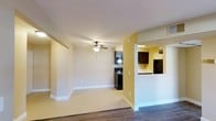 This single-floor residence offers an open and functional layout designed for comfortable everyday living. The main living area is finished with light-toned tile flooring and features a ceiling fan with a built-in light fixture, providing both air circulation and ambient lighting. Neutral, cream-colored walls are complemented by warm yellow accent walls that add a cheerful, inviting energy throughout the space.

The kitchen is partially open to the living area via a half-wall pass-through, creating a connected yet defined cooking space. It is equipped with dark-toned cabinetry, a black range with overhead microwave, and additional upper cabinetry for generous storage. The contrast between the dark cabinetry and the warm yellow walls gives the kitchen a bold, contemporary character.

The transition between the living area and kitchen is marked by a shift from tile to hardwood-style flooring, subtly delineating each zone while maintaining a cohesive flow throughout the home. White baseboards throughout the space provide a clean, polished finish. The overall design balances practicality with warm aesthetic choices, making this single-level home feel welcoming, well-organized, and move-in ready.
