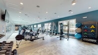 This single-story fitness center is thoughtfully designed to accommodate a comprehensive range of workout needs across two distinct zones. The main exercise area is spacious and open, featuring patterned light-toned tile flooring that extends throughout. Bold teal accent walls contrast with crisp white ceilings outfitted with recessed lighting and ceiling fans, creating an energizing yet comfortable atmosphere. A full wall of floor-to-ceiling mirrors amplifies the sense of space while allowing users to monitor their form during workouts.

The strength training section is well-stocked with cable machines, a multi-function weight rack, free weights including a complete dumbbell rack, stability balls, medicine balls, and resistance accessories neatly organized on dedicated storage racks. A separate cardio zone houses multiple treadmills and stair climbers, positioned alongside expansive floor-to-ceiling glass sliding doors that provide direct views of an outdoor pool and palm trees, flooding the room with natural light. An adjacent cycling studio offers a more intimate setting with its own teal accent wall, hardwood-style flooring, and dedicated spin bikes. Together, these areas deliver a cohesive, fully functional fitness environment.