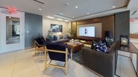 This well-appointed single-floor amenity space offers a diverse range of functional areas united by a cohesive modern aesthetic. Large-format white tile flooring flows throughout, providing a clean, polished foundation across every zone. The central lounge area is elegantly furnished with dark velvet and gold-accented seating arranged around a large-screen display, complemented by recessed tray ceilings, warm lighting, and curated artwork that adds a sophisticated touch. Adjacent to the lounge, a games room features a full-size billiards table beneath a statement chandelier, a bar-height counter with stools, and its own wall-mounted display. The conference area presents a spacious white table surrounded by upholstered chairs, set atop a patterned area rug, with greenery and large-scale wall art enhancing the professional atmosphere. The business center offers a dedicated row of desktop workstations along a clean white desk, set against a bold teal accent wall. A sleek kitchen and dining area rounds out the space, with warm wood cabinetry, a white island with gold-framed barstools, a professional coffee station, and sculptural pendant lighting. A built-in display niche with glass shelving adds a refined architectural detail near the entryway.