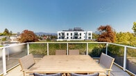Located in the vibrant Kensington-Cedar Cottage neighbourhood of Vancouver, BC, this rooftop terrace at 1408 E 15th Ave offers an exceptional outdoor living experience elevated above the surrounding streetscape. The terrace is finished with large-format stone pavers that provide a clean, contemporary aesthetic underfoot, while a sleek white-framed glass railing system ensures unobstructed sightlines in every direction.

The outdoor space is generously sized, comfortably accommodating a full teak dining table and set of woven rattan chairs, making it well-suited for al fresco entertaining. A lush green hedge wall lines one side of the terrace, adding a layer of natural privacy and vibrant colour to the space. Access is provided via French doors set into the building's clean, modern facade.

The views from the terrace are a true highlight: to the north, the North Shore mountains rise prominently against clear skies, while the surrounding East Vancouver neighbourhood stretches out below, dotted with mature trees including striking Japanese maples. A neighbouring contemporary white building frames part of the view, reinforcing the urban character of the location. The terrace receives abundant natural light throughout the day, making it an ideal retreat for enjoying Vancouver's sunny seasons in a private, elevated setting.