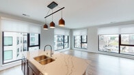 This well-appointed single-floor residence offers a thoughtfully designed open-concept layout that seamlessly connects the kitchen, dining, and living areas. The kitchen is a standout feature, showcasing rich dark wood cabinetry, quartz countertops with elegant veining, stainless steel appliances including a refrigerator, range, and built-in microwave, and a central island with a double stainless steel sink topped by stylish pendant lighting. Light wood-tone flooring runs throughout the main living areas, complementing the neutral color palette and enhancing the bright, airy atmosphere created by an array of large windows fitted with roller shades.

The living area is generously proportioned, bathed in natural light from multiple oversized windows that offer views of the surrounding neighborhood. Two bedrooms feature plush carpeting, large windows, and ample closet space, with the primary bedroom also providing direct access to a private balcony through sliding glass doors.

Both bathrooms are finished with the same quartz countertops found in the kitchen, dark wood vanities, undermount sinks, and wood-look flooring. Each bathroom includes a bathtub and shower combination. The overall design is clean, modern, and cohesive, creating a comfortable and functional living environment.