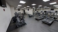 This fitness center is a single-floor facility designed to accommodate a comprehensive workout experience. The space is generously sized, offering ample room for movement between equipment stations. The flooring consists of durable, dark rubberized material that provides both traction and impact absorption, ideal for high-traffic fitness use.

The room is well-lit by a grid of recessed fluorescent panel lights across the drop ceiling, creating a bright and evenly illuminated environment throughout. A full-length mirror runs along one wall, visually expanding the space while allowing users to monitor their form during exercise.

The cardio section is prominently featured, housing multiple treadmills, elliptical trainers, and stationary bikes arranged for easy access and spacing. A cable and strength training machine station is visible toward the rear, rounding out the facility's functional range. Wall-mounted flat-screen televisions are positioned at multiple points around the room, providing entertainment options during workouts.

The overall aesthetic is clean and utilitarian, with neutral white walls and a practical layout that prioritizes functionality. The space is well-organized and maintained, making it a capable facility for both cardio and resistance training.
