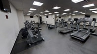 This fitness center is a single-floor facility designed to accommodate a comprehensive workout experience. The space is generously sized, offering ample room for movement between equipment stations. The flooring consists of durable, dark rubberized material that provides both traction and impact absorption, ideal for high-traffic fitness use.

The room is well-lit by a grid of recessed fluorescent panel lights across the drop ceiling, creating a bright and evenly illuminated environment throughout. A full-length mirror runs along one wall, visually expanding the space while allowing users to monitor their form during exercise.

The cardio section is prominently featured, housing multiple treadmills, elliptical trainers, and stationary bikes arranged for easy access and spacing. A cable and strength training machine station is visible toward the rear, rounding out the facility's functional range. Wall-mounted flat-screen televisions are positioned at multiple points around the room, providing entertainment options during workouts.

The overall aesthetic is clean and utilitarian, with neutral white walls and a practical layout that prioritizes functionality. The space is well-organized and maintained, making it a capable facility for both cardio and resistance training.