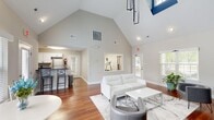 Located at 2401 Fieldsway Dr in Chesapeake, Virginia, this single-floor community space is thoughtfully designed to offer both comfort and functionality. The open-concept layout is anchored by dramatic vaulted ceilings that lend an impressive sense of height and airiness throughout the main living area. Rich hardwood flooring flows seamlessly across the entire space, adding warmth and visual continuity.

The living area is a welcoming focal point, highlighted by a marble-surround fireplace set against a bold blue accent wall — a striking design detail that adds character and depth. Multiple windows fitted with plantation shutters invite abundant natural light, while providing views of the surrounding greenery. A large window pass-through connects the living area to a fitness room, maintaining visual openness.

A cozy dining nook sits adjacent to the living space, featuring a window-facing table ideal for casual seating. The kitchen is well-appointed with crisp white cabinetry, dark granite countertops, stainless steel appliances, and a breakfast bar with seating for two — combining practicality with clean, modern aesthetics.

Throughout the space, a neutral color palette of whites and soft grays is complemented by carefully chosen accent tones, creating a polished and inviting atmosphere suited for community gatherings or everyday use.