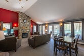 This single-floor space is defined by its striking vaulted ceilings, which soar above an open-concept living and dining area to create an airy, expansive feel. The focal point of the living room is a floor-to-ceiling stacked stone fireplace set against a bold red accent wall, lending the space a warm, lodge-inspired character. A ceiling fan is mounted at the peak of the vaulted ceiling, and wall sconces provide ambient lighting throughout. The living area is furnished with plush sofas and armchairs arranged around a round coffee table, with a television and media console positioned nearby. Soft carpeting runs throughout the main living and dining spaces.

The dining area accommodates a large dark wood table with seating for several guests and is positioned adjacent to large windows dressed in gray curtains, allowing ample natural light to filter in. French doors provide additional brightness and access to the outdoors. The kitchen, accessible directly from the dining area, features wood cabinetry, a refrigerator, and standard appliances. A hallway leads from the dining space to additional rooms. The overall ambiance is cozy and inviting, blending rustic and casual elements effectively.