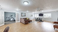Located at 75 N Orchard Dr in Warsaw, Indiana, this single-floor space offers a thoughtfully organized layout comprising a generous open living area and a dedicated exercise room. The living area is a standout, featuring warm light hardwood flooring, soft gray walls, and a decorative embossed ceiling that adds a touch of character throughout. Crown molding frames the space cleanly, while a ceiling fan and multiple flush-mount light fixtures provide comfortable illumination. The area is divided into distinct functional zones: a comfortable lounge space anchored by a sectional sofa, a dining area with a wall-mounted TV and ample natural light from multiple windows, and a work nook with desk setup. A kitchen is accessible from the living area, featuring wood cabinetry and standard appliances. The exercise room is accessed via a classic French door and is carpeted for comfort, housing a variety of fitness equipment including a stationary bike, elliptical, treadmill, and free weights, along with a wall-mounted TV and a water cooler. A second room also features additional exercise equipment and its own exterior door for convenient access. Natural light filters throughout the space, creating a bright, welcoming atmosphere suited for both productivity and active living.