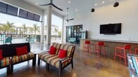 This single-story amenity lounge is designed to seamlessly blend indoor comfort with outdoor living. The space is anchored by soaring ceilings and polished, variegated concrete floors that extend throughout, lending an industrial-chic character to the overall aesthetic. Expansive floor-to-ceiling black-framed glass walls and sliding doors connect the interior to a covered outdoor patio, creating a fluid transition between the two areas and allowing generous natural light to pour in.

The interior lounge area features multiple seating arrangements with bold, multicolored striped sofas and chairs accented by vivid red throw pillows, adding a lively, energetic atmosphere. A built-in wet bar with a corrugated metal front runs along one wall, topped with a sleek counter and accompanied by bright red bar stools. A wall-mounted flat-screen television is positioned above the bar for entertainment.

The covered outdoor patio mirrors the interior's energy, offering additional lounge seating and a oversized Connect Four game, perfect for recreational use. Views from the space overlook a resort-style pool area lined with palm trees. Ceiling fans are installed both indoors and on the covered patio, ensuring comfort throughout. The cohesive design and vibrant furnishings make this a dynamic, social gathering space.