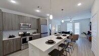 This contemporary single-floor residence showcases a thoughtfully designed open-concept layout that seamlessly connects the kitchen, dining, and living areas. The kitchen is a standout feature, equipped with stainless steel appliances — including a refrigerator, electric range, and built-in microwave — complemented by dark wood cabinetry, quartz countertops, and a large waterfall island with an undermount sink and bar seating. Pendant lighting accents the island, adding a modern touch. The living area is bright and airy, benefiting from large windows and a ceiling fan, with light wood-style flooring flowing throughout the main spaces. The bedrooms are well-proportioned and filled with natural light from wide windows, each featuring ceiling fans and light-toned flooring. The primary bathroom includes a single-vanity with dark wood cabinetry, a bathtub with shower, and direct access to a generously sized walk-in closet outfitted with built-in shelving and hanging rods on multiple sides. The overall aesthetic is clean and contemporary, with white walls, modern fixtures, and an open, inviting atmosphere that ties each area of the residence together cohesively.