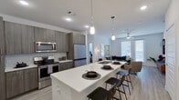 This contemporary single-floor residence showcases a thoughtfully designed open-concept layout that seamlessly connects the kitchen, dining, and living areas. The kitchen is a standout feature, equipped with stainless steel appliances — including a refrigerator, electric range, and built-in microwave — complemented by dark wood cabinetry, quartz countertops, and a large waterfall island with an undermount sink and bar seating. Pendant lighting accents the island, adding a modern touch. The living area is bright and airy, benefiting from large windows and a ceiling fan, with light wood-style flooring flowing throughout the main spaces. The bedrooms are well-proportioned and filled with natural light from wide windows, each featuring ceiling fans and light-toned flooring. The primary bathroom includes a single-vanity with dark wood cabinetry, a bathtub with shower, and direct access to a generously sized walk-in closet outfitted with built-in shelving and hanging rods on multiple sides. The overall aesthetic is clean and contemporary, with white walls, modern fixtures, and an open, inviting atmosphere that ties each area of the residence together cohesively.