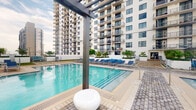 This outdoor amenity space is defined by a generously sized swimming pool with clear, blue-tinted water and a tiled surround designed for comfortable poolside use. A striking dark metal pergola structure anchors the foreground of the pool deck, adding an architectural focal point to the open-air environment. The pool area is lined with rows of blue lounge chairs, providing ample seating for relaxation throughout the day.

The space is framed on multiple sides by a tall, modern residential tower with a white facade, large balconies, and grid-pattern windows that rise prominently above the pool level. Tropical plantings, including palm trees and low greenery, are integrated along the perimeter, softening the built environment and reinforcing a resort-like ambiance. The pool deck itself features patterned paving with drainage channels, contributing to both functionality and visual order.

Open to natural light and the surrounding sky, the area benefits from an airy, sun-filled atmosphere well-suited for outdoor leisure. The overall design reflects a contemporary urban aesthetic, combining clean architectural lines with amenity-focused outdoor living.