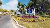 This vibrant apartment community boasts an array of impressive amenities. The centerpiece is a spacious, resort-style pool surrounded by paved decking and lush landscaping. Residents can relax in comfortable lounge areas featuring red umbrellas and modern outdoor furniture. A children's playground and outdoor gaming area with foosball add to the family-friendly atmosphere. The property's entrance is adorned with a beautifully landscaped sign and tropical plants. Well-maintained buildings with blue siding are nestled among mature trees, creating a serene environment. Additional conveniences include on-site car care facilities. The overall design seamlessly blends Florida's natural beauty with modern, comfortable living spaces.