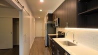 This single-floor space showcases a well-designed interior with a contemporary aesthetic. The galley-style kitchen is a standout feature, equipped with dark espresso-toned cabinetry that extends to the ceiling, maximizing vertical storage. Light quartz countertops provide a crisp contrast to the rich cabinetry, while a classic subway tile backsplash adds texture and visual interest. Stainless steel appliances, including a refrigerator, electric range, built-in microwave, and a deep undermount sink with a modern arc-neck faucet, complete the functional and polished kitchen setup. Recessed lighting illuminates the space efficiently, highlighting the warm-toned hardwood-style flooring that runs throughout. A sliding barn door is visible along the hallway, adding a modern architectural detail while saving floor space. The corridor leads toward the entry door, giving the layout a clean, linear flow. White walls and ceilings keep the interior bright and open, complementing the darker cabinetry and flooring for a balanced, modern contrast throughout the unit.