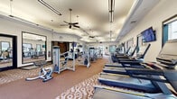 Located at 1 Garden Park Blvd in Cherry Hill, NJ, this fitness center offers a spacious, thoughtfully designed workout environment spanning a single floor. The main exercise area is generously proportioned, providing ample room for a wide array of cardio machines, including treadmills and ellipticals, alongside an extensive selection of strength-training equipment from brands such as Atlantis. Dedicated free weight stations, weight racks, and multi-use cable machines are well-distributed throughout the space, ensuring a comprehensive workout experience.

The interior features warm neutral-toned walls, elevated tray ceilings with ceiling fans for air circulation, and a combination of pendant lighting and linear overhead fixtures that create a bright, inviting atmosphere. Patterned carpeting in the main workout zones adds visual interest, while large framed mirrors along the walls enhance the sense of openness. Multiple windows and glass doors allow natural light to filter in and offer views of the surrounding greenery outdoors.

A separate entry corridor provides a polished transition into the facility, complete with tile flooring, wall-mounted water fountains, restroom access, and decorative artwork. Mounted flat-screen televisions are positioned throughout the workout floor for convenience, contributing to a modern, well-appointed fitness environment.