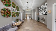 This single-floor community clubhouse is designed with a contemporary aesthetic and thoughtfully curated amenities throughout. The grand entrance lobby sets a refined tone, featuring large-format stone tile flooring, elevated tray ceilings with crown molding, and a striking geometric chandelier. One wall is adorned with decorative living moss and wood art installations, while bold black-and-white graphic artwork anchors the opposite side. Built-in water stations and a seating area with a rustic wood coffee table provide functional comfort for visitors.

Moving deeper into the space, a spacious lounge area offers hardwood flooring, a tiled fireplace wall with a mounted television, and plush seating arrangements. Tall transom windows and French doors flood the room with natural light and provide views to an outdoor terrace. An arched window adds architectural charm at the far end.

A curved kitchen bar serves as a social hub, complete with a marble-style countertop, mosaic tile backsplash, stainless steel range hood, and a dramatic cascade of globe pendant lights overhead. Chrome barstools line the counter, creating an inviting gathering spot.

A glass-enclosed co-working room offers a bright, focused work environment with views to the exterior. A cozy gallery nook with curated artwork and intimate seating rounds out this well-appointed community facility.