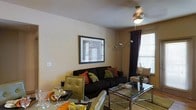 This single-floor space offers a comfortable and stylishly appointed interior that blends functionality with inviting design. The living area is anchored by a dark sofa adorned with vibrant orange and green patterned throw pillows, creating a bold contrast against the warm, neutral-toned walls. A ceiling fan with an integrated light fixture provides both comfort and ambient illumination, while a sleek arc floor lamp adds supplemental lighting and a modern touch. Natural light filters in through a window dressed with white horizontal blinds and framed by rich, dark curtains, lending a warm glow to the room. A framed artwork piece on the wall adds a decorative focal point, contributing to the curated aesthetic of the space. The glass-top coffee table with a chrome frame sits at the center of the seating area, complemented by additional seating and a side chair nearby. The dining area, visible in the foreground, features a glass dining table with a chrome base and olive-toned upholstered chairs, set with decorative napkins and glassware. The tiled flooring ties the spaces together seamlessly, creating a cohesive and welcoming atmosphere throughout this well-designed single-floor home.