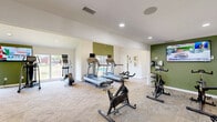 Located at 610 Paradise Ct in Greenwood, Indiana, this fitness center offers a well-appointed exercise space designed to support a comprehensive workout routine. The single-floor facility is thoughtfully laid out with distinct zones for cardio, strength training, and cycling. Vaulted ceilings fitted with ceiling fans enhance the sense of openness and airflow throughout, while recessed lighting ensures the space remains bright and functional at any hour. Large windows along the perimeter flood the room with natural light and provide views of the surrounding outdoor pool area and greenery, creating an energizing atmosphere. The flooring features durable, neutral-toned carpet tile suited for high-traffic fitness use. Cardio equipment includes multiple treadmills and elliptical trainers, while a dedicated cycling area houses stationary spin bikes alongside a wall-mounted display screen. A multi-station strength machine and additional resistance equipment serve those focused on muscle conditioning. Accent walls in bold olive green add a fresh, modern aesthetic contrast to the crisp white walls and trim. Practical amenities such as sanitation stations, a waste receptacle, and posted gym rules contribute to a clean and organized environment. Glass doors connect to an adjacent space, allowing for easy access and a sense of continuity within the broader facility.