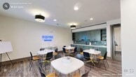 Located at 152 Market St in Pittsboro, NC, Inspire by Liberty – Briar Chapel is a thoughtfully designed single-floor studio space that blends functional amenities with a modern, welcoming aesthetic. The open-concept layout accommodates both social dining and creative activities with ease.

The kitchen and dining area is anchored by a sleek, full-length countertop finished in crisp white quartz, complemented by sage-green shaker-style cabinetry and a striking teal subway tile backsplash. A deep undermount sink with a matte black faucet adds a refined, contemporary touch. Upper cabinets with open shelving provide both storage and display space. Multiple round white dining tables paired with a mix of navy and wood-toned chairs create a casual yet stylish gathering environment, all illuminated by recessed lighting and drum pendant fixtures.

The adjacent activity room is highlighted by a glowing "INSPIRE – Briar Chapel" neon sign that serves as a vibrant focal point. This versatile space features the same wood-look flooring that flows throughout, and is equipped with mobile storage carts and a sewing machine, making it well-suited for crafting workshops or group projects. Large windows along one wall bathe the space in natural light while offering views of the surrounding outdoor greenery, contributing to an overall atmosphere that is bright, energetic, and creatively inspiring.