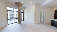 This thoughtfully designed single-floor space seamlessly blends industrial character with contemporary finishes. At its centerpiece stands a striking, weathered industrial column with a distinctive flared base, serving as a bold architectural focal point that pays homage to the building's heritage. Light-toned hardwood flooring extends throughout, lending warmth and a sense of openness to the interior.

The main living area is bathed in natural light, courtesy of expansive floor-to-ceiling black-framed glass doors that lead directly to an outdoor balcony, offering views of the surrounding urban landscape. The crisp white walls and recessed ceiling lighting contribute to a clean, bright ambiance that feels both airy and welcoming.

A partially visible adjacent room, accessible through a white-paneled door, hints at additional functional spaces within the layout. The contrast between the raw, patinated industrial column and the refined, minimalist surroundings creates a dynamic visual tension that defines the character of the space. Overall, the design strikes a compelling balance between historic industrial elements and modern residential sensibility, making it a distinctive and versatile environment.