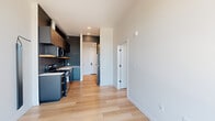 Located at 1714 NW Northrup St in Portland, Oregon, this efficiently designed one-bedroom, one-bathroom unit delivers a modern urban living experience. Light-toned hardwood flooring runs throughout the entire space, creating a cohesive and clean aesthetic from room to room.

The kitchen is a standout feature, outfitted with dark navy cabinetry, a white quartz countertop, stainless steel appliances including an electric range and over-the-range microwave, and an open wood floating shelf that adds a warm contrast to the contemporary palette. A hallway connects the kitchen and living area to the private spaces beyond.

The bedroom is a bright, serene retreat benefiting from large floor-to-ceiling windows that frame views of the surrounding Portland cityscape, filling the room with abundant natural light. A dedicated closet provides organized storage adjacent to the bedroom.

The bathroom is finished with clean, modern details including light wood cabinetry with a warm walnut tone, a backlit vanity mirror, matte black fixtures, white subway tile in the shower/tub surround, and matching light hardwood flooring. Crisp white walls throughout the unit enhance the sense of openness and keep the design feeling fresh and contemporary.