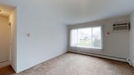 This single-floor space presents a clean, minimalist living area characterized by its neutral palette and straightforward layout. The room features smooth white walls and a beige wall-to-wall carpet that adds warmth underfoot while maintaining a simple, understated aesthetic. A large picture window dressed with vertical blinds serves as the focal point of the main living area, allowing an ample amount of natural light to fill the space and offering views of a green outdoor area and neighboring structures. The window's generous size contributes to an open, airy atmosphere throughout the room. A baseboard heating unit runs beneath the window, providing an efficient and unobtrusive source of warmth. The white ceiling is smooth and uniform, complementing the clean lines of the walls. The overall design of the space is practical and functional, with an open floor plan that allows for flexible furniture arrangements. The neutral tones throughout create a blank canvas well-suited for a variety of interior design preferences.