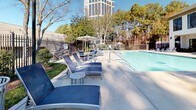 This outdoor amenity space centers around a generously sized swimming pool with clear, blue water that reflects the open sky above. The expansive concrete pool deck provides ample room for relaxation and socializing, lined with blue sling-style lounge chairs and side tables arranged for comfort and convenience. Multiple umbrella-shaded seating areas offer relief from the sun, with additional table-and-chair groupings ideal for gathering. The pool area is fully enclosed by a decorative iron fence, providing both security and an open, airy aesthetic. Lush landscaping, including mature trees, manicured shrubs, and planted beds, frames the perimeter and adds a natural, tranquil character to the setting. A prominent multi-story clubhouse structure with large windows, a covered balcony, and clean architectural lines overlooks one end of the pool, lending an upscale, resort-like atmosphere. Tall pine trees and a modern high-rise building visible beyond the fencing provide a distinctive urban-meets-nature backdrop. Stairways connect tiered levels of the deck, leading to additional seating areas and a pergola. The overall ambiance is bright, open, and inviting, well-suited for both leisurely lounging and social gatherings in an outdoor setting.