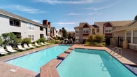 Cheyenne Crossing is a well-appointed residential community designed to offer a range of outdoor and recreational amenities. The centerpiece of the community is a generously sized outdoor swimming pool, finished with classic brick tile edging and surrounded by a stamped concrete deck that provides ample space for lounging and socializing. A row of poolside lounge chairs lines the adjacent lawn area, offering a relaxing retreat under open skies. Complementing the main pool is a connected spa, detailed with deep blue tile accents.

The pool area also features a covered patio space with a gated iron entry, white support columns, and seating that includes a circular outdoor dining set — ideal for outdoor gatherings. A dedicated bike share station, branded as Cheyenne Crossing Bikeshare, is conveniently located poolside, encouraging an active lifestyle. A separate building houses the Fitness Center, accessible via a gated entrance with ornate ironwork and a welcoming covered porch with bench seating.

The surrounding residential buildings feature a traditional architectural style with gabled rooflines, cedar-toned and neutral siding, prominent chimneys, and exterior staircases. Mature trees and maintained shrubbery frame the property, contributing to a green and comfortable atmosphere throughout the community.