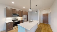 This thoughtfully designed single-floor residence showcases a contemporary open-concept layout with a seamless flow between the kitchen and living areas. The kitchen is a standout feature, equipped with rich dark walnut-toned cabinetry, stainless steel appliances including a refrigerator, range, and microwave, and a generous white quartz island with an undermount sink. Stylish pendant lights hang above the island, adding character and warmth to the space. Light hardwood-style flooring runs throughout the main living areas, complementing the clean white walls and bright, airy atmosphere. The living area opens directly to a private balcony through large sliding glass doors framed in black, offering pleasant outdoor views of greenery and water. Additional rooms include well-proportioned bedrooms, one featuring a large picture window with black framing that frames scenic outdoor views, and soft carpet underfoot. The bathroom is clean and functional, with light cabinetry, a white quartz vanity, and a framed mirror. Stone-accented balcony columns add an architectural detail visible from inside. A dedicated utility and laundry room houses a stacked Whirlpool washer and dryer alongside the building's mechanical systems, providing practical convenience entirely within the unit.