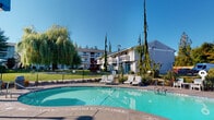 This outdoor recreational area centers around a generously sized swimming pool with crystal-clear turquoise water, set within a thoughtfully designed pool deck finished in stamped concrete. The pool's freeform shape accommodates a range of swimmers, with clearly marked depth indicators along its edges. Surrounding the pool is a spacious deck furnished with multiple reclining lounge chairs and patio seating, including umbrella-shaded tables that provide relief from the sun on warm days. The area is fully enclosed by a wrought-iron security fence with gated access, ensuring a safe and private environment for users. Lush, mature landscaping frames the space, including graceful weeping willow trees, tall columnar evergreens, ornamental grasses, and colorful flowering shrubs that add natural beauty and a sense of seclusion. A street lamp positioned within the enclosure suggests the area is usable into the evening hours. The backdrop features a multi-story residential building with balconies and well-maintained green lawns, contributing to the community atmosphere. Safety signage is posted throughout, including no-diving and swim-at-your-own-risk notices. The overall setting is bright, open, and inviting, ideal for relaxation and recreation.