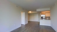 This single-floor space showcases a clean, contemporary design centered around an open-concept living and kitchen area. The main living space is finished with rich, dark hardwood flooring that runs throughout, creating a cohesive and polished aesthetic. Neutral-toned walls and a smooth white ceiling contribute to a bright, airy atmosphere, while multiple HVAC vents indicate a well-integrated climate control system.

The kitchen is positioned along one side of the open layout and features warm wood-toned cabinetry paired with a stainless steel refrigerator, offering a modern contrast to the neutral surroundings. A white kitchen island or peninsula provides additional workspace and serves as a natural divider between the kitchen and the living area.

Two white-paneled doors are visible from the main living space, suggesting access to additional rooms such as bedrooms or utility areas. The layout appears thoughtfully arranged, allowing for a generous flow between zones. The combination of practical finishes, open sightlines, and functional design elements makes this a versatile and inviting space suited to a variety of living arrangements.