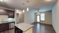 This single-story residence showcases a thoughtfully designed open-concept layout that seamlessly connects the kitchen and living areas. The kitchen is a standout feature, equipped with rich dark espresso cabinetry extending to the ceiling, granite countertops, and a full suite of stainless steel appliances, including a refrigerator with ice maker, electric range, microwave, and dishwasher. A central island with a matching granite surface provides additional prep and seating space, while pendant lighting and a track light fixture illuminate the area effectively. The living space extends naturally from the kitchen, finished with warm wood-tone flooring throughout and a ceiling fan with integrated lighting. Windows fitted with blinds allow natural light to filter in while maintaining privacy. The bathroom is well-appointed, featuring a dual-sink vanity topped with granite, dark cabinetry with brushed nickel hardware, a large mirror, and ample overhead lighting. A tub and shower combination with neutral tile surround completes the bathroom. The overall aesthetic is cohesive, combining dark wood tones, granite surfaces, and neutral walls for a clean, contemporary feel.