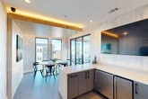 Located at 1401 Third Ave S in Birmingham, Alabama, this stylish kitchen and dining space delivers a refined, contemporary aesthetic with thoughtful architectural detailing throughout. The open layout seamlessly connects a sleek kitchen to a dining area, all unified by large-format tile flooring in a cool gray tone that grounds the space with a clean, modern feel.

The kitchen features an L-shaped white countertop paired with matte charcoal cabinetry accented by black hardware, creating a striking contrast. A black undermount sink with a minimalist faucet adds a bold focal point, while stainless steel appliances complete the functional design. Warm ambient lighting from LED strips beneath a natural wood ceiling beam introduces an inviting warmth that balances the cooler palette of the walls, which display a subtle textured finish throughout.

The dining area is anchored by a marble-topped table surrounded by contemporary black chairs, positioned to take full advantage of the floor-to-ceiling black-framed sliding glass doors. These doors open to an outdoor terrace, flooding the interior with natural light and offering expansive views of the Birmingham skyline. A wall-mounted television and curated decorative accents, including driftwood sculptures and a large-scale abstract artwork, add personality and polish to this elevated, design-forward space.