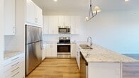 This well-appointed single-floor unit offers a clean, contemporary design with a thoughtful layout that maximizes both comfort and functionality. The open-concept kitchen is a standout feature, boasting crisp white shaker-style cabinetry, a classic subway tile backsplash, and striking granite countertops with natural veining. A suite of stainless steel appliances — including a refrigerator, electric range, microwave, and dishwasher — completes the space, while a modern geometric pendant light fixture adds a stylish touch overhead. The kitchen flows seamlessly into the living area, which is finished with light hardwood-style flooring that extends throughout the main living spaces, lending warmth and continuity to the design. Large windows in the living area and bedroom invite abundant natural light and provide elevated views of the surrounding neighborhood. The bedroom features plush carpeting, a built-in closet with shelving, and a bright window that enhances the airy feel of the room. The bathroom is clean and functional, offering a tub-shower combination, a white vanity with drawer storage, and wood-tone flooring. Throughout the unit, neutral tones, recessed lighting, and a fresh, move-in-ready finish create a bright and inviting atmosphere.