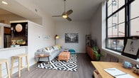 This well-appointed single-floor residence showcases a modern, loft-inspired design characterized by soaring ceilings and expansive industrial-style grid windows that flood the space with abundant natural light and offer open views of the surrounding urban streetscape. Throughout the main living areas, durable hardwood-style flooring provides a cohesive and contemporary aesthetic.

The spacious living area is anchored by a ceiling fan and comfortably accommodates a full seating arrangement, with the large windows serving as a dramatic backdrop. Adjacent to the living space, a dedicated workspace with direct window access creates an ideal environment for productivity. The kitchen features warm wood cabinetry, clean white countertops, stainless steel appliances including a full-size refrigerator, and a breakfast bar with seating — combining functionality with style.

The bedroom is a cozy, well-defined retreat with a distinctive teal accent wall, built-in closet storage with open shelving, and plush carpeting underfoot. A teal entry accent wall adds a bold, welcoming pop of color to the corridor. The bathroom is finished with clean tile flooring, a sleek dark vanity with a white countertop, a large mirror, and contemporary fixtures. Overall, the space strikes a balance between urban modern style and comfortable, practical living.