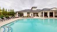 Located at 601 McCaw Bridge Street in Raleigh, NC, this single-story outdoor amenity complex offers a resort-inspired experience designed for relaxation and socializing. At the heart of the space is a generously sized swimming pool with a smooth, freeform shape, finished with a vivid turquoise interior and framed by a broad, light-toned tile pool deck. Ample lounge chairs are arranged throughout the deck, providing dedicated spaces for sunbathing and leisure.

The centerpiece building features a classic white board-and-batten farmhouse exterior with a dark shingled gable roof and a distinctive cupola. A covered veranda runs along the poolside facade, supported by crisp white columns and equipped with ceiling fans, offering a shaded seating and dining area that opens through large glass doors to the interior. A wrought-iron railing encloses the veranda's poolside edge, creating a defined yet open transition between indoor and outdoor spaces.

Beyond the pool, the grounds include a landscaped lawn area anchored by a circular fire pit surrounded by seating, ideal for evening gatherings. A separate wood-framed pergola provides additional shaded seating with outdoor furniture. Lush greenery and manicured shrubs border the perimeter, enhancing the sense of privacy and natural ambiance throughout the complex.