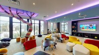 This lively children's activity space is thoughtfully designed to inspire creativity and play at every turn. The open-plan layout is anchored by a striking multicolored sculptural tree column that serves as a bold centerpiece, drawing the eye upward toward stylishly recessed ceilings accented with ambient purple and teal lighting. Floor-to-ceiling windows line multiple walls, flooding the space with natural light and offering views of lush palm trees and the surrounding neighborhood beyond.

The room is organized into distinct activity zones, each tailored to different interests and age groups. A cozy media corner features a large flat-screen TV mounted above built-in shelving stocked with books, board games, and toys, complemented by playful curved seating in vibrant red and yellow. A dedicated learning and computer station lines one wall, furnished with child-sized desks and chairs, while a central activity area offers whimsically shaped tables and bunny-eared chairs for arts, crafts, and group play.

A standout butterfly mural wall adds an artistic focal point, while soft carpet tiles provide a safe and comfortable surface throughout. The overall ambiance is energetic yet welcoming, blending thoughtful design with child-friendly functionality to create an inviting environment for young residents and visitors alike.