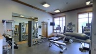 This fitness center is a thoughtfully designed, single-floor facility built to accommodate a full range of exercise needs. The space features a clean, functional layout with neutral-toned walls accented by warm wood crown molding and trim, lending a polished yet welcoming atmosphere. Flush-mounted ceiling light panels provide bright, even illumination throughout the room, complementing the natural light that filters in through a window fitted with horizontal blinds. The flooring consists of durable, dark rubber matting well-suited for high-traffic workout use. A large wall-mounted mirror along one side of the room visually expands the space and allows users to monitor their form during exercise. The equipment selection is comprehensive, including a multi-station cable and weight machine, an adjustable workout bench, dumbbells, a rowing machine, treadmills, and additional cardio equipment. A wall-mounted television is positioned for easy viewing during cardio sessions. Signage identifying the space as a Fitness Center is clearly visible, and the overall layout allows for comfortable movement between stations. The combination of strength and cardio offerings, thoughtful lighting, and quality finishes makes this a capable and inviting facility for daily exercise.